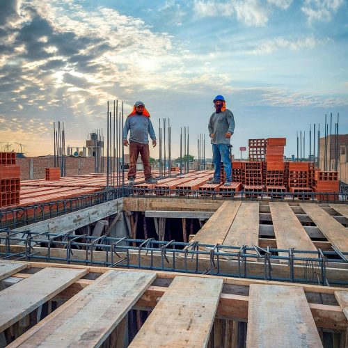 Two construction workers stand on a building site under a cloud-filled sky. Sunlight illuminates the scene.