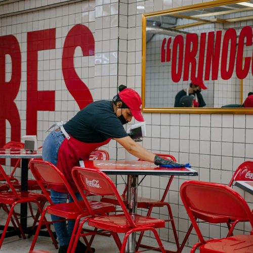 A worker cleans tables in a restaurant, emphasizing hygiene and safety protocols.