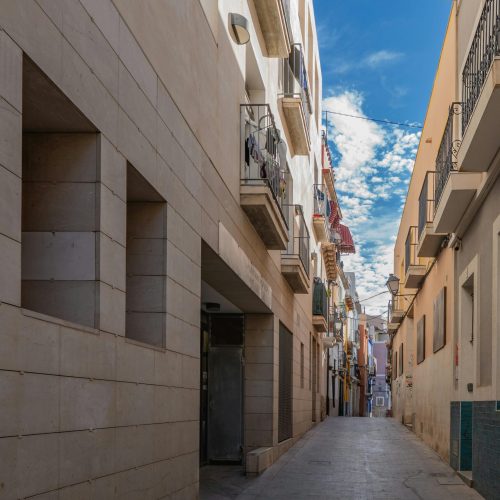 A scenic view of a narrow street with colorful buildings in Alicante, Spain under a clear blue sky.