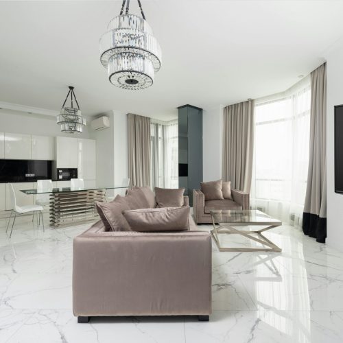 Interior of modern light studio with couch and armchair with pillows near glass table and chairs under chandelier next to open kitchen with white cabinets and windows with curtains