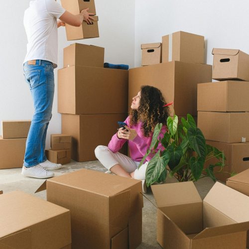 Side view of young male in casual clothes arranging stack of cardboard boxes during relocation into new apartment with girlfriend sitting on floor and using smartphone